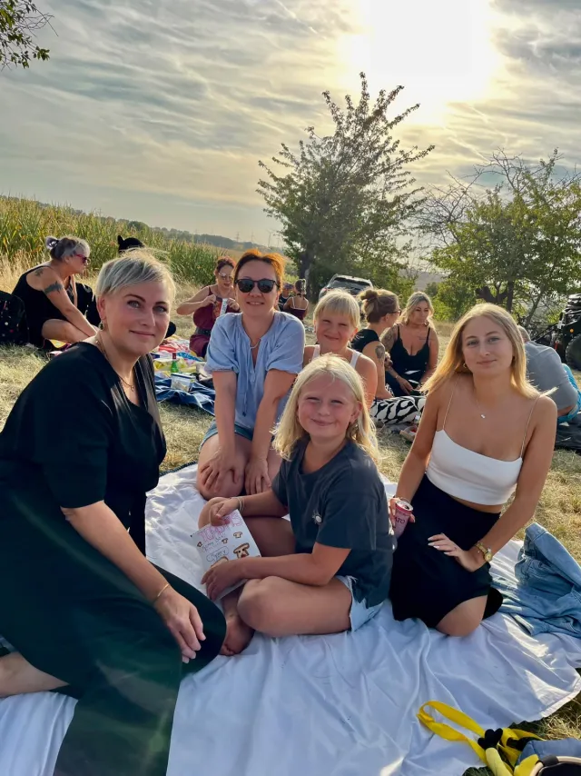 Eine Gruppe sitzt auf einer weißen Picknickdecke im Gras, dahinter ein Maisfeld und weitere Besucher; die tiefstehende Sonne scheint durch dünne Wolken.