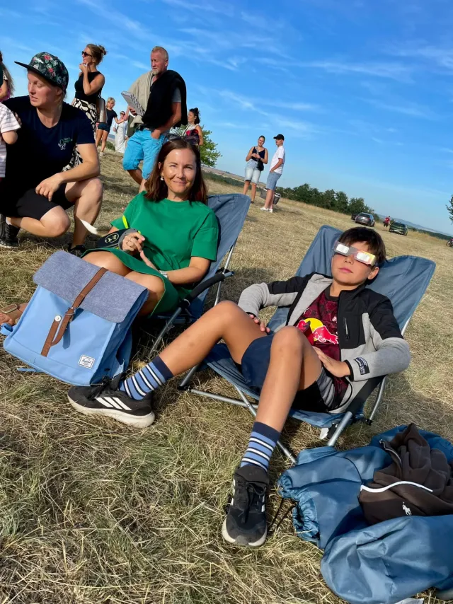 Zwei Gäste liegen nebeneinander in Campingstühlen auf der Wiese, einer davon mit Sonnenfinsternis-Schutzbrille, und schauen in den Himmel.