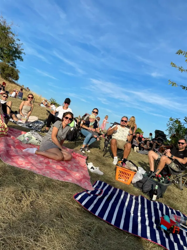 Blick über die Picknickwiese am Hang: viele Menschen auf Decken und in Campingstühlen unter blauem Himmel, im Vordergrund eine rot geblümte und eine blau-weiß gestreifte Decke.