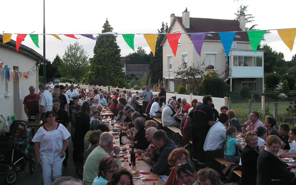 Eine lange Tafel zieht sich durch eine Dorfstraße: Menschen sitzen dicht beieinander beim Essen, über ihnen hängen bunte Wimpelketten zwischen den Häusern.