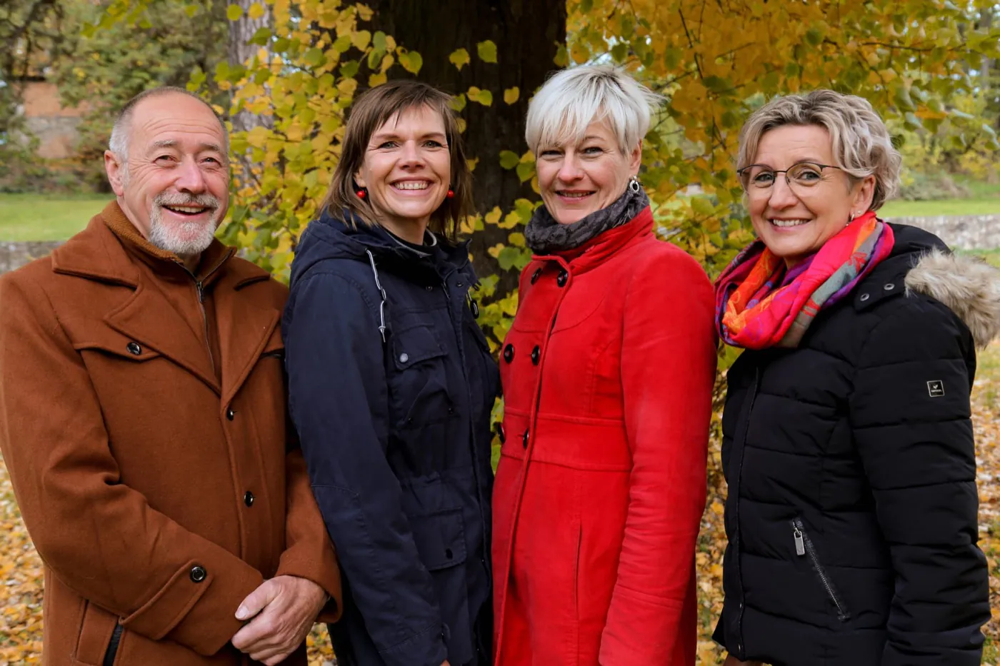Die vier Vorstandsmitglieder der KulturWerkstatt stehen dicht nebeneinander vor einem Baumstamm im herbstlich gelben Laub und schauen lächelnd in die Kamera.